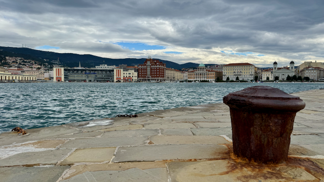 View across the waterfront in Trieste, Italy, with the stone pier of Molo Audace in the foreground and a weathered cast-iron mooring bollard overlooking the Gulf of Trieste. Across the water, the city’s historic waterfront, including Piazza Unità d’Italia and its elegant neoclassical buildings, stretches beneath dark, dramatic storm clouds, creating a striking contrast between the restless sea and the timeless architecture.
